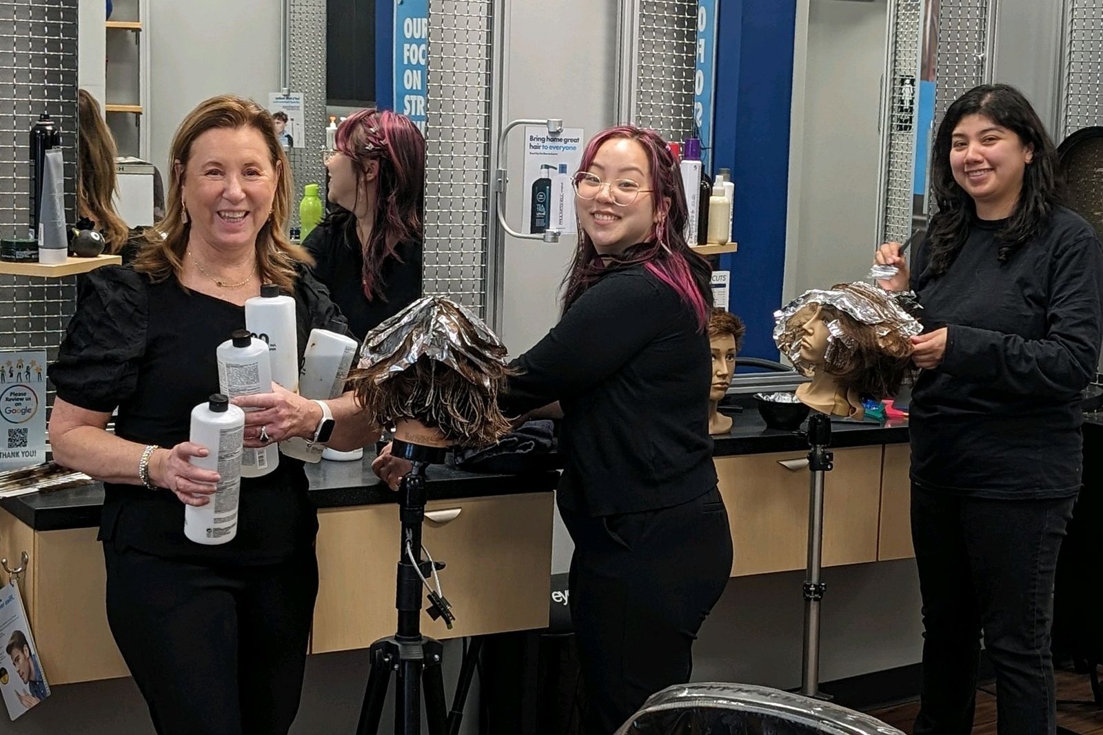 An educator and two stylists smiling during foil highlighting practice on mannequin heads in one of our salons